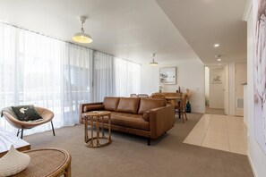 A modern living room with a brown leather sofa, a round wicker chair, and a dining table with chairs. - Luxury Apartment w/ Pool, Parking, Walk to Beach (Forster)