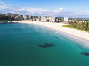 A coastal area with a sandy beach and a clear blue sea. There are several buildings in the background, likely hotels or resorts. - Luxury Apartment w/ Pool, Parking, Walk to Beach (Forster)