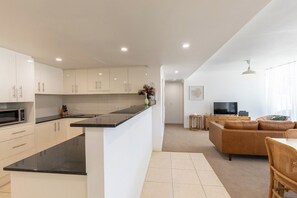 A modern kitchen with white cabinets and a black countertop, a living area with a brown leather sofa and a TV, and a dining area with a table and chairs. - Luxury Apartment w/ Pool, Parking, Walk to Beach (Forster)