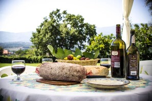 Een buitentafel gedekt met een fles wijn, een brood, een kaaswiel, een mand met fruit en een glas rode wijn.