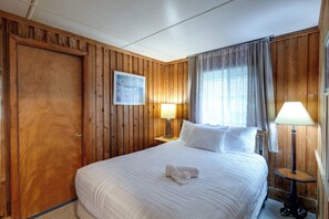 A hotel room with a wooden paneled wall, a bed with white bedding, two bedside lamps, and a window with curtains. - Flip Flop Cottage: Lake Access in Clermont (Clermont)