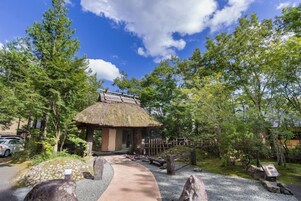 A traditional Japanese-style building with a thatched roof, surrounded by a gravel path and greenery.