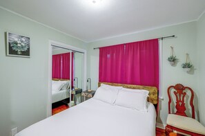 Ein Schlafzimmer mit einem Bett, einem Spiegel, einem Fenster mit Vorhängen, einem Nachttisch, einem Sessel und einer an der Wand befestigten Pflanze.