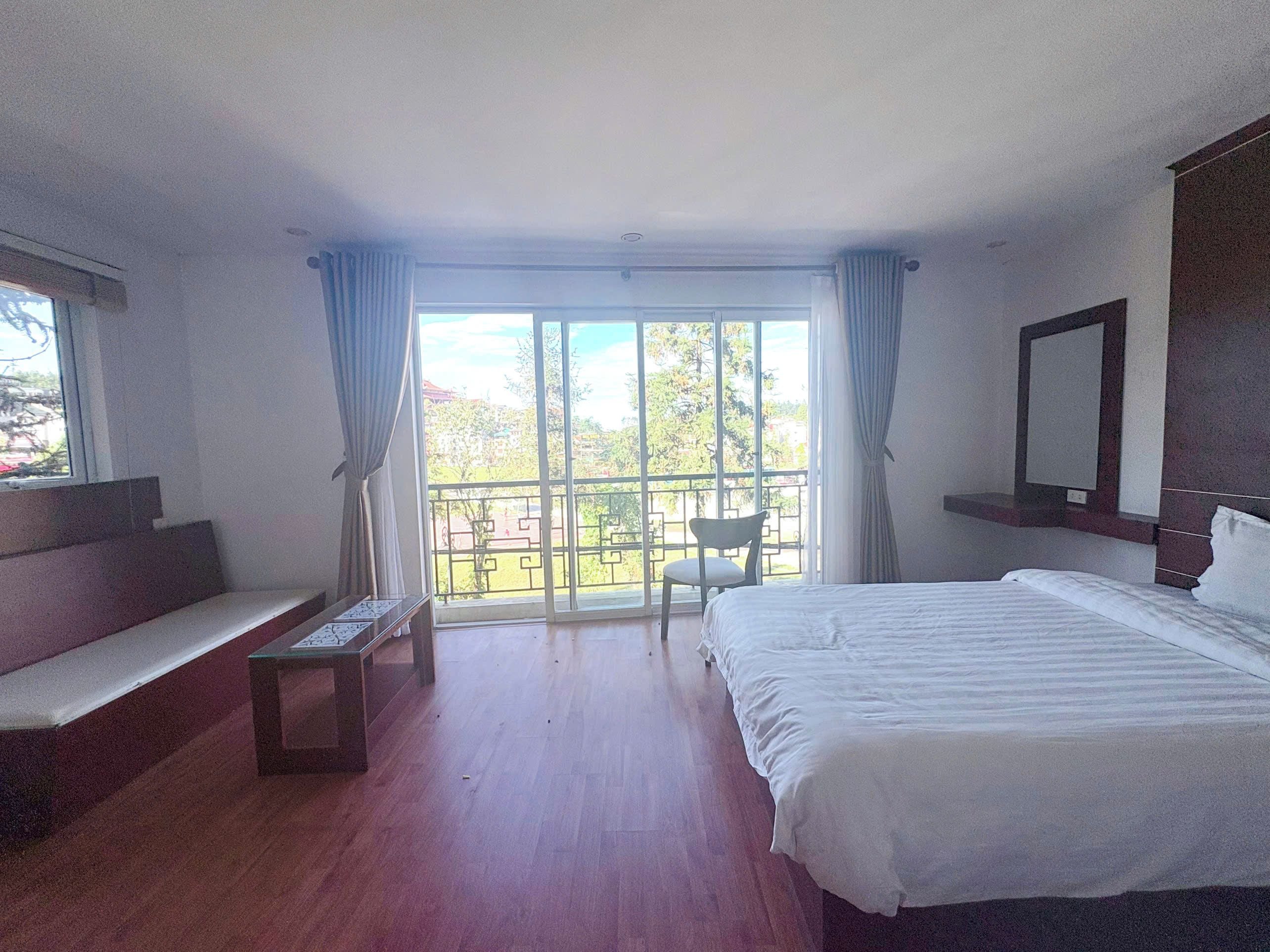 Une chambre d’hôtel avec un lit, une petite table, une chaise, un balcon donnant sur un espace verdoyant et une fenêtre avec des rideaux.
