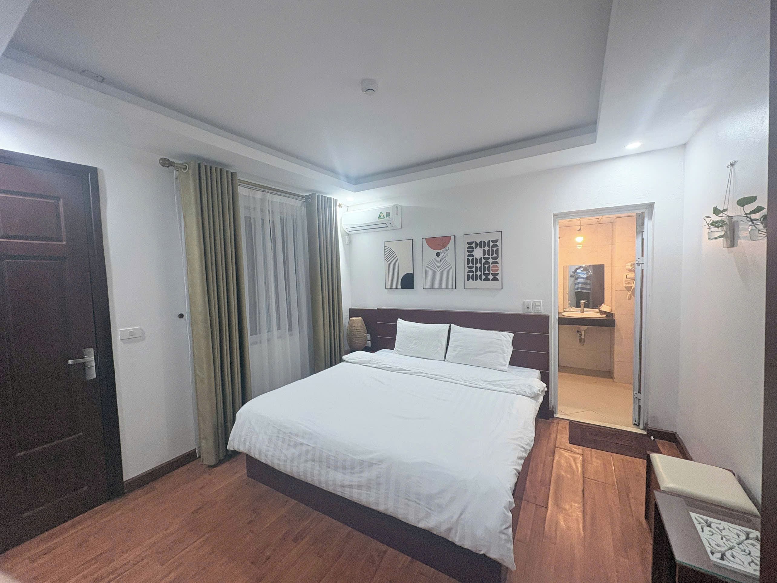 Une chambre d’hôtel avec un lit, une table de chevet, un miroir, une porte et une salle de bain visible à travers une porte ouverte.