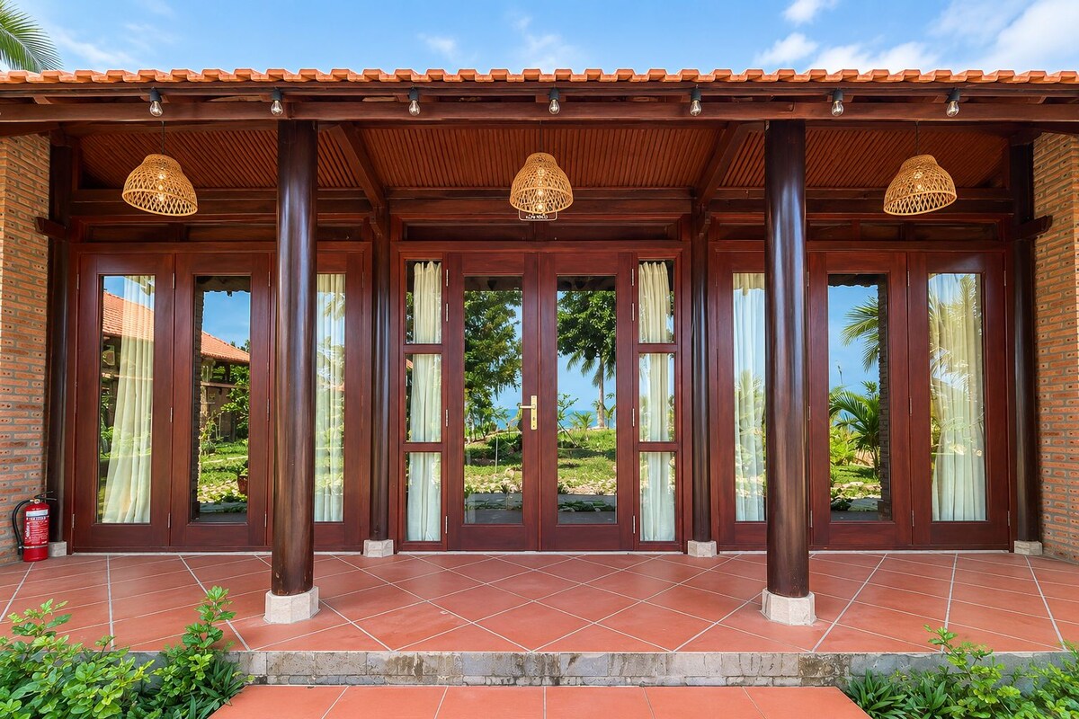 Uma área externa coberta, com pilares de madeira, grandes janelas e piso de azulejos.