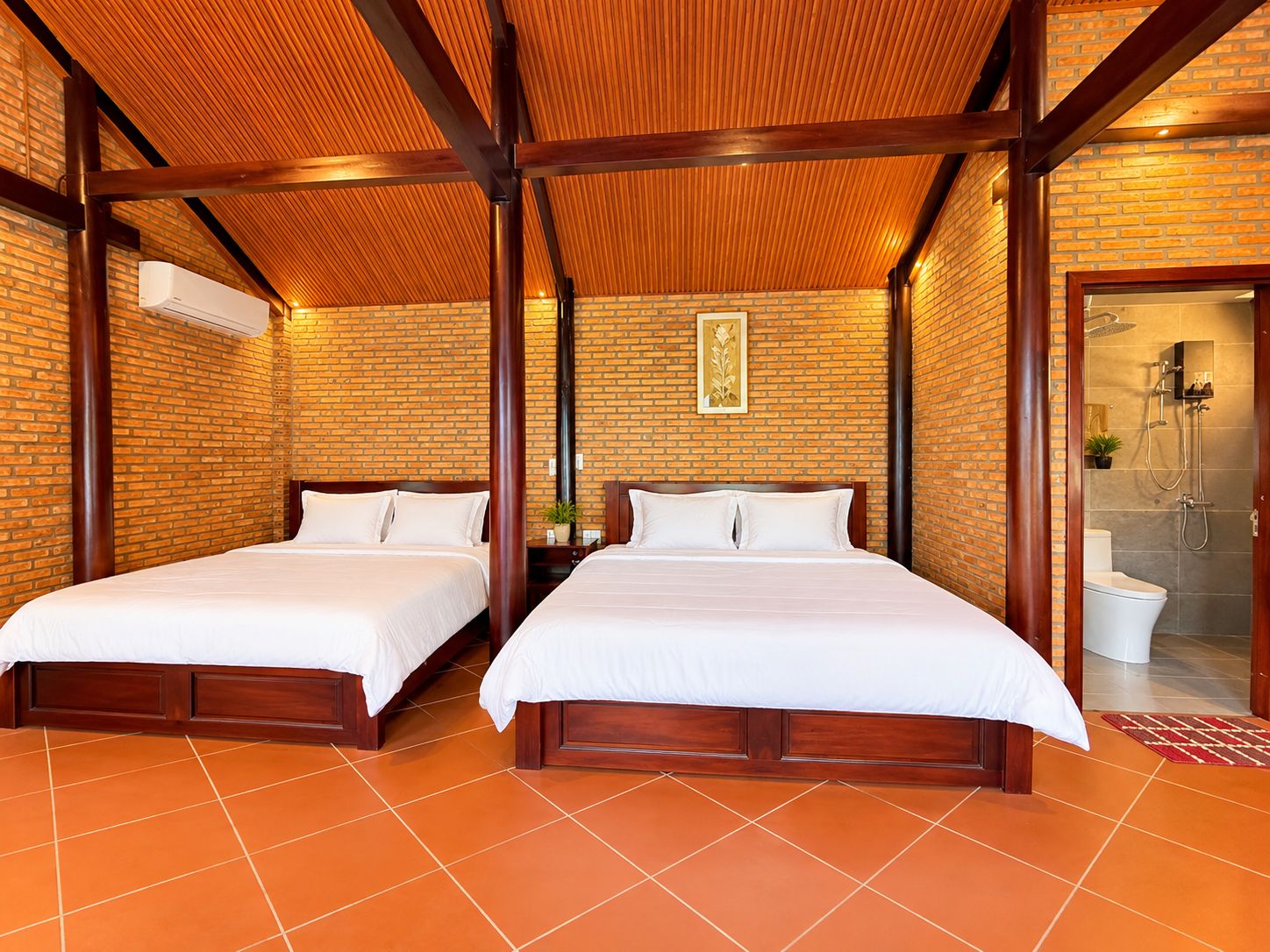 A hotel room with two beds, a brick wall, a wooden ceiling, and a bathroom visible through an open door.