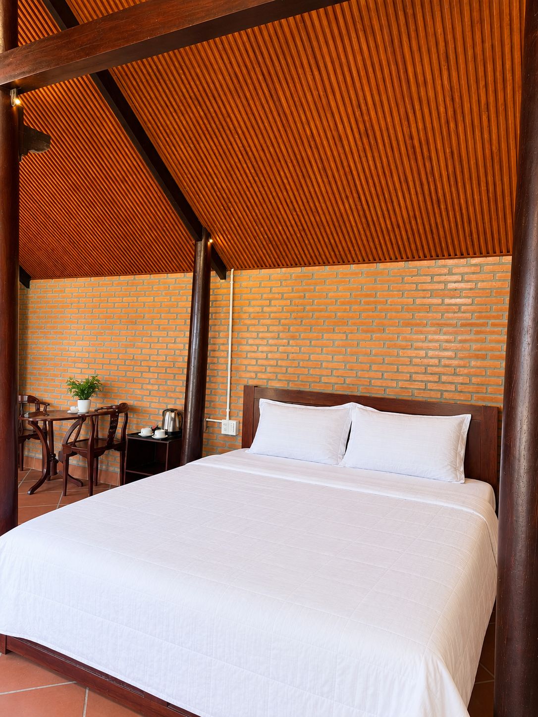 Een hotelkamer met een bakstenen muur, een houten plafond, een bed met wit beddengoed, een klein tafeltje met een plant en een stoel.