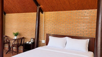 Een hotelkamer met een bakstenen muur, een houten plafond, een bed met wit beddengoed, een klein tafeltje met een plant en een stoel.