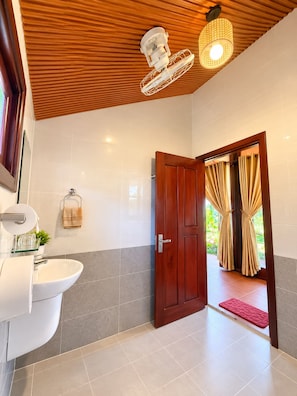 Casa de banho com teto de madeira, uma pia branca e porta que dá para uma varanda com cortinas.