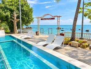 Uma área à beira da piscina com espreguiçadeiras, uma placa indicando 'Dragon Beach Resort' e vista para o oceano.