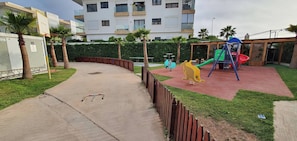 A playground with a slide and swings, surrounded by a low fence and palm trees. - 3 Bedrooms Pools Terrace Beach Dar Bouazza (Dar Bouazza)