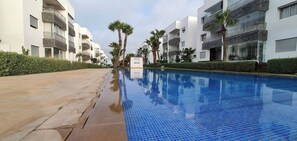 A swimming pool with a tiled edge, surrounded by modern buildings with balconies and palm trees. - 3 Bedrooms Pools Terrace Beach Dar Bouazza (Dar Bouazza)