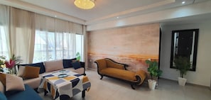 A modern living room with a sofa, armchair, and coffee table. There is a large window with curtains, a chandelier, and potted plants. - 3 Bedrooms Pools Terrace Beach Dar Bouazza (Dar Bouazza)