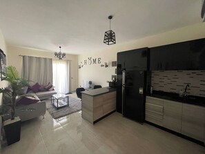 Eine moderne Küche mit einem schwarzen Kühlschrank, ein Essbereich mit einem Glastisch und ein Wohnzimmer mit einer Couch und einer Pflanze.