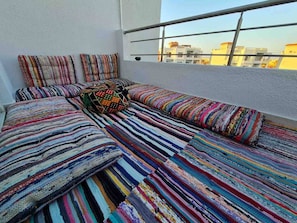 A balcony with a sofa and pillows, a patterned rug, and a view of buildings. - Sunshine & Smiles – Nabq’s Most Lovely Apartment (South Sinai Governorate)