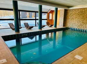 Une piscine intérieure offrant une vue dégagée sur l’extérieur, des chaises longues en bois et un mur en pierre.