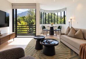 A modern living room with a sofa, coffee table, and TV. There is a balcony with a view of greenery and mountains. - Modern luxury Claremont apartment - sleeps 6 (Cape Town)