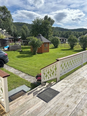 Eine Holzterrasse mit weißem Gittergeländer, eine kleine Holzhütte und ein Rasenstück mit Bäumen.
