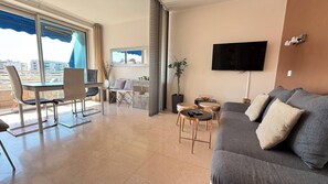 A modern living room with a grey sofa, a TV mounted on the wall, a dining area with a table and chairs, and a balcony with a view of the city. - Beautiful 2 bedroom terrace and parking (Cannes)