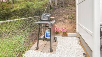 Ein Grill mit Propangasflasche, umgeben von einem Maschendrahtzaun, Kies und rosa Blüten.