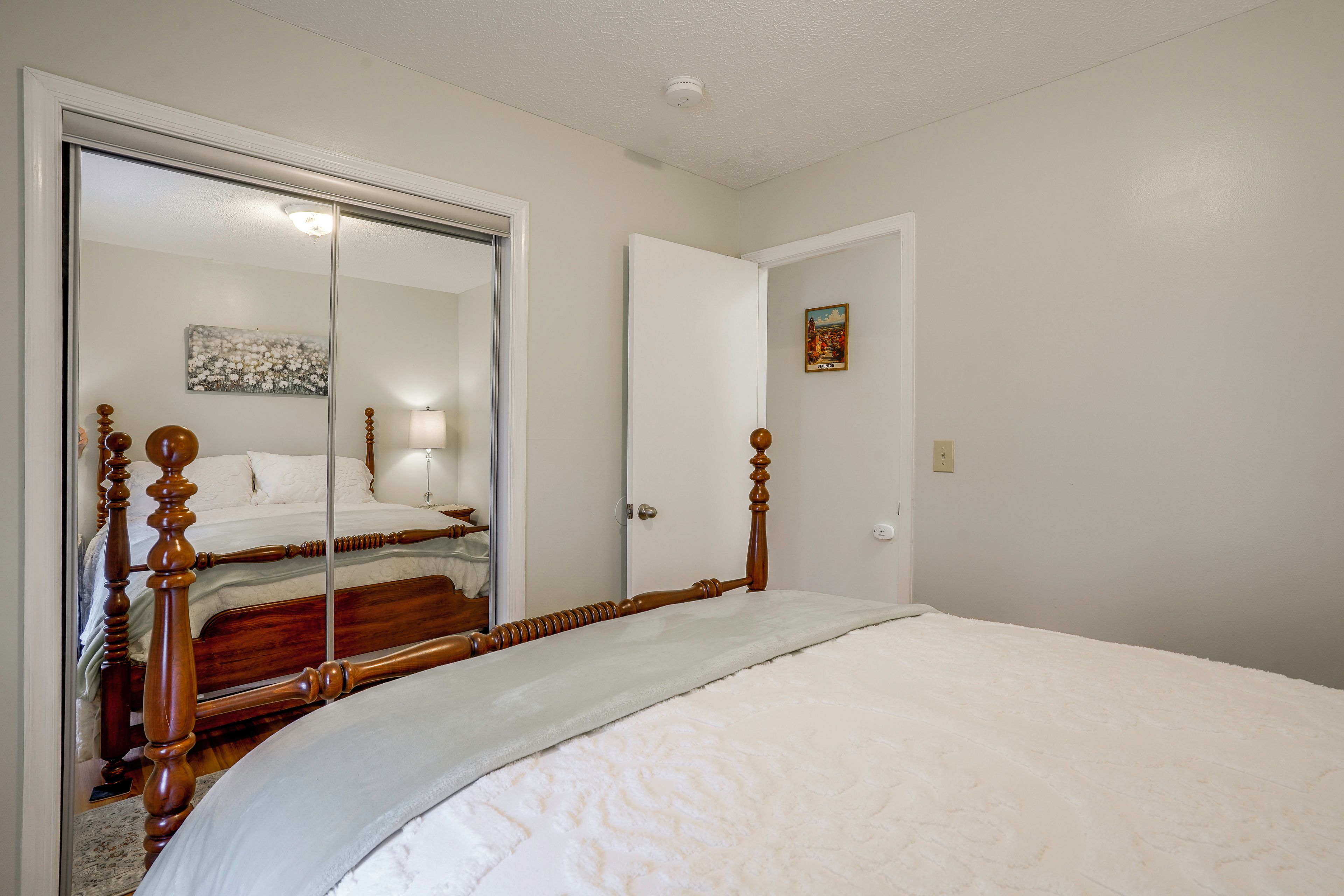 A bedroom with a large mirror, a bed with wooden headboard, a door, and a wall-mounted light fixture.