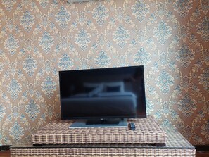 A television on a stand with a remote control beside it, set against a patterned wallpaper. - 591 DSA by Hostech.ai (Shah Alam)
