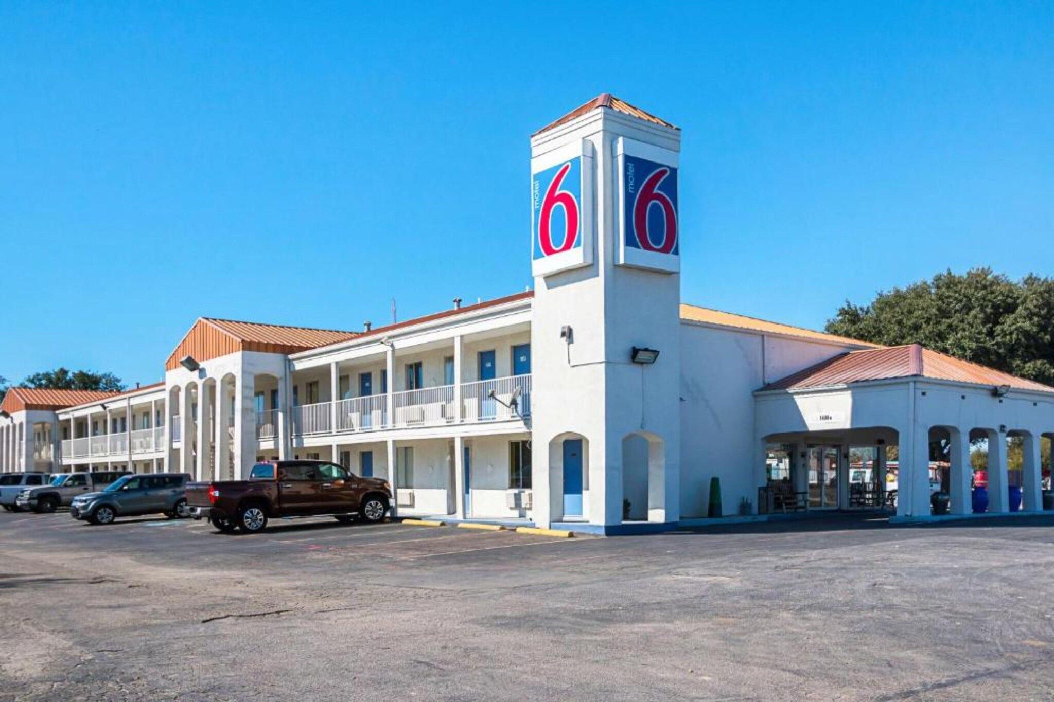 A motel with a central tower featuring the number '6', a parking lot, and a building with a red roof.