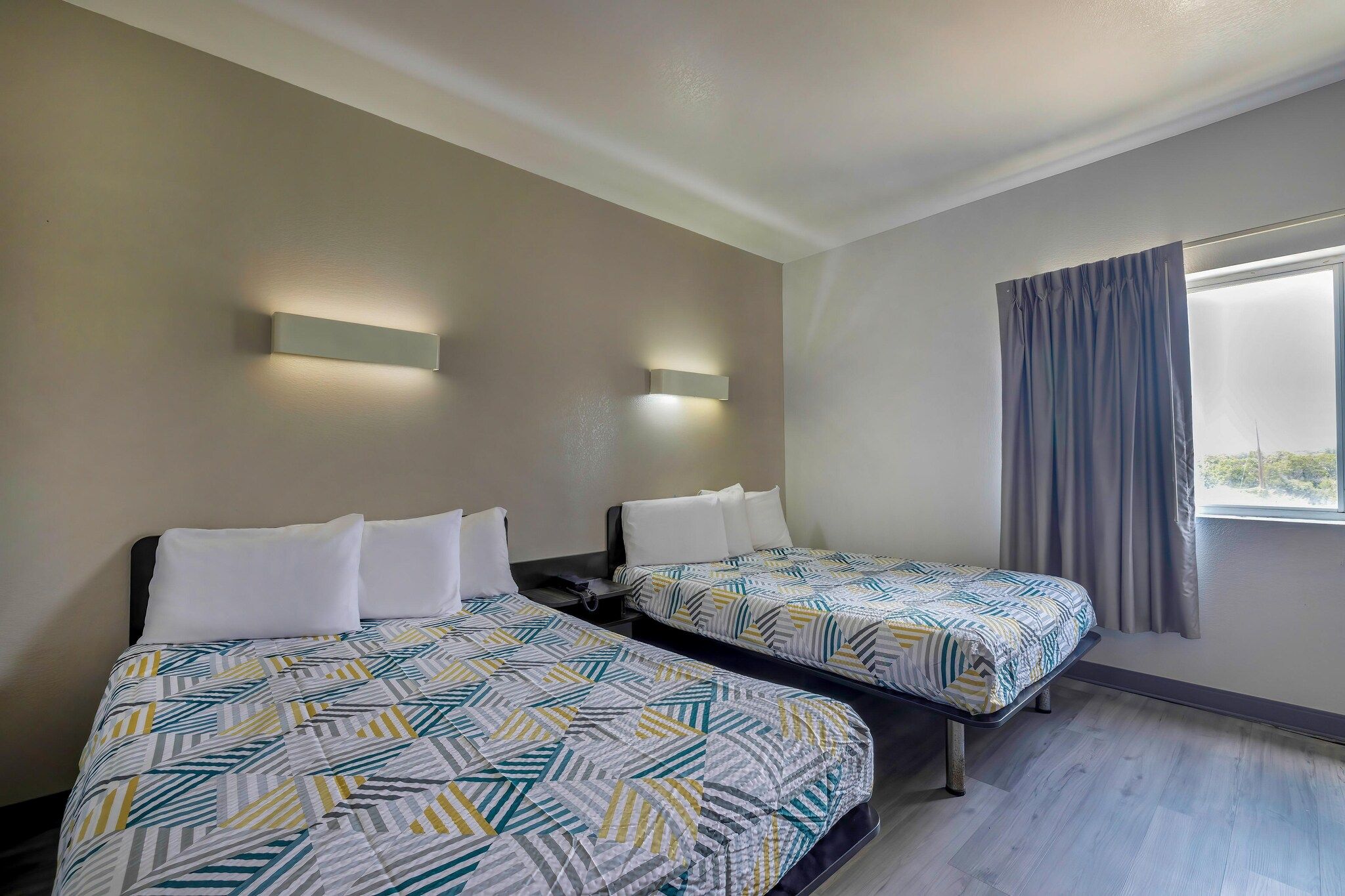 A hotel room with two beds, a window with curtains, and wall-mounted lighting.