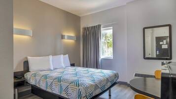 A hotel room with a bed, bedside table, lamp, window with curtains, and a mirror on the wall.