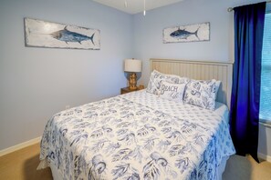 Ein Schlafzimmer mit einem Bett, einem Nachttisch, einer Lampe, zwei Wandbildern und einem Fenster mit Vorhängen.