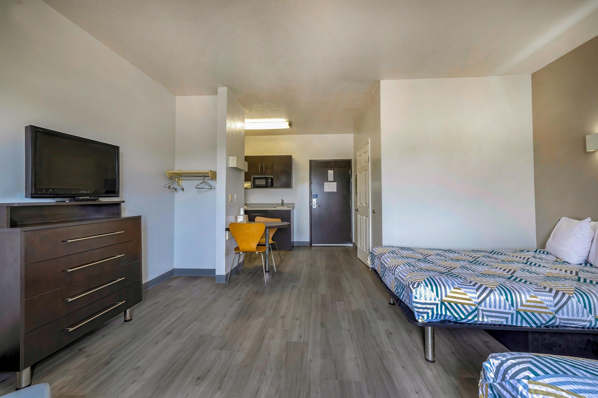 A hotel room with a TV, a desk with a chair, a bed with patterned bedding, and a kitchenette.