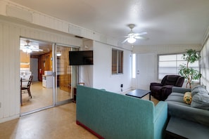 Ein Zimmer mit einer Schiebetür zu einer Küche, einem Deckenventilator, einem flachbildfernseher an der Wand und einer Sitzecke mit einer Couch und Sesseln.