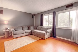 Ein Wohnzimmer mit einem Ecksofa, einem kleinen Beistelltisch, einer Lampe und einem großen Fenster mit Vorhängen.