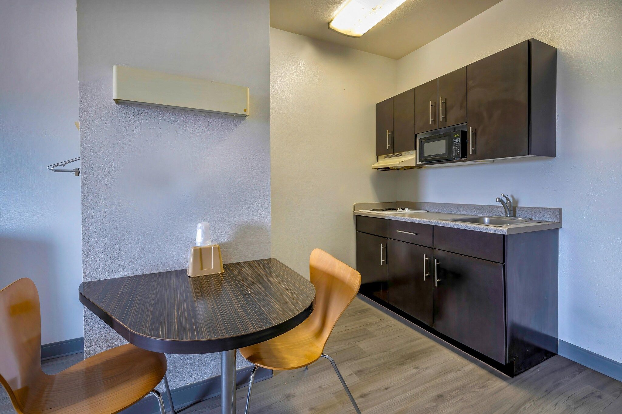 Eine kompakte Küche mit Mikrowelle, Spüle und dunklen Schränken. Ein kleiner Tisch mit Stühlen und eine Flasche stehen darauf.