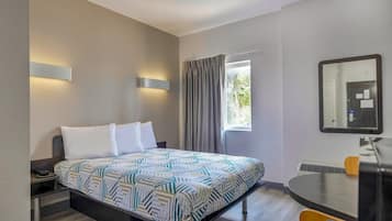 A hotel room with a bed, bedside table, lamp, window with curtains, and a mirror on the wall.
