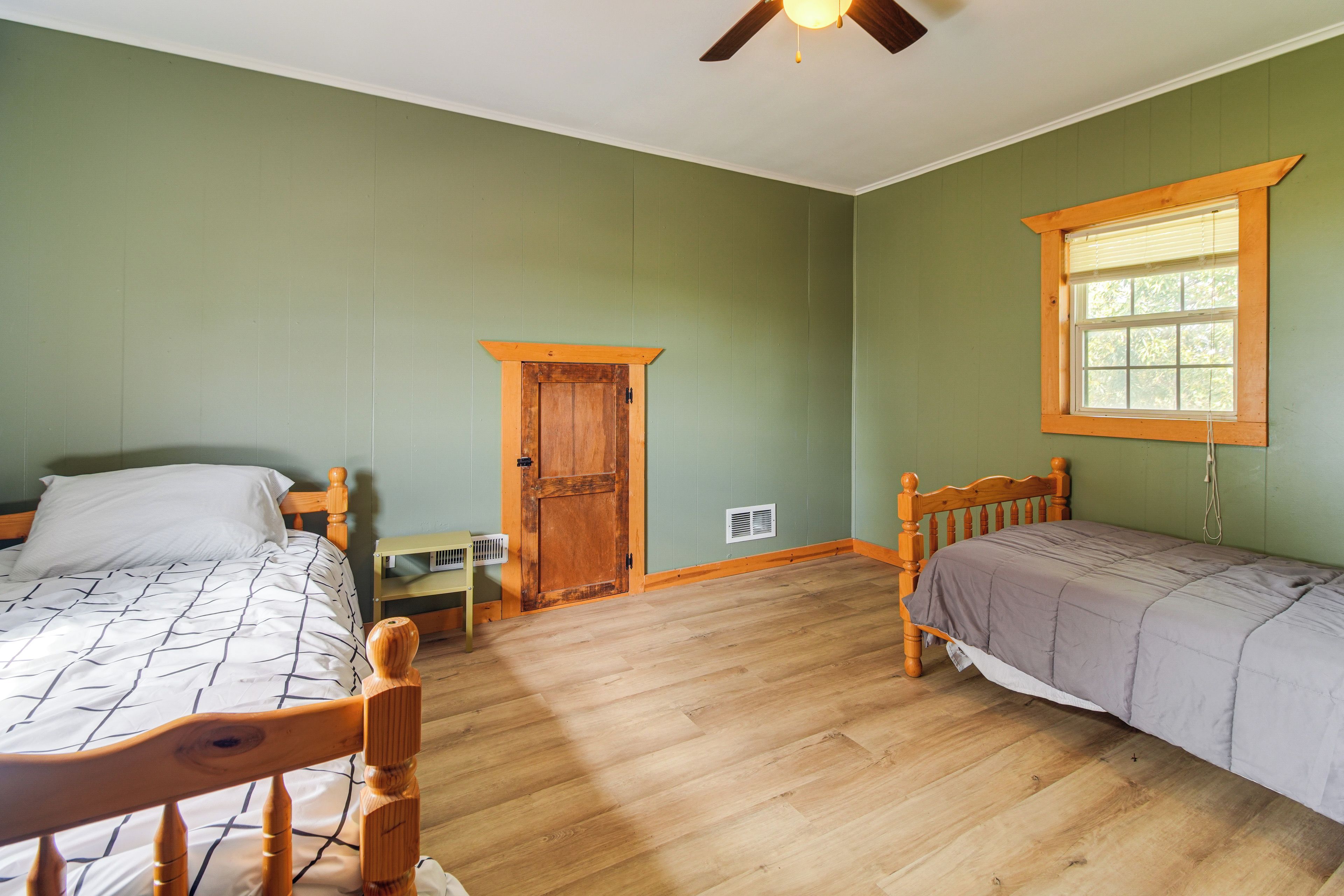 A room with two wooden beds, a window with blinds, a door, a radiator, and a ceiling fan.