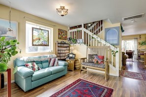 A living room with a blue sofa, a bookshelf, a staircase, and a large window. - Creekside New Castle Townhome w/ Scenic Deck (New Castle)