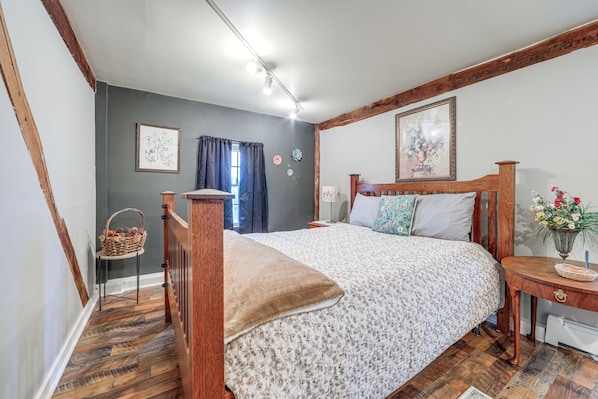 A bedroom with a wooden bed, a nightstand, a bedside table, a window with curtains, and a wall with a framed picture.