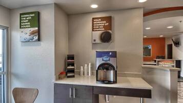 Zona de recepción de hotel con una máquina de café, un cartel que invita a descargar la aplicación y un anuncio que indica que el cargo matutino es gratuito.