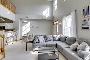 Ein modernes Wohnzimmer mit einem grauen Ecksofa, einem Glastisch und einem Essbereich mit Stühlen und einem kleinen Tisch.