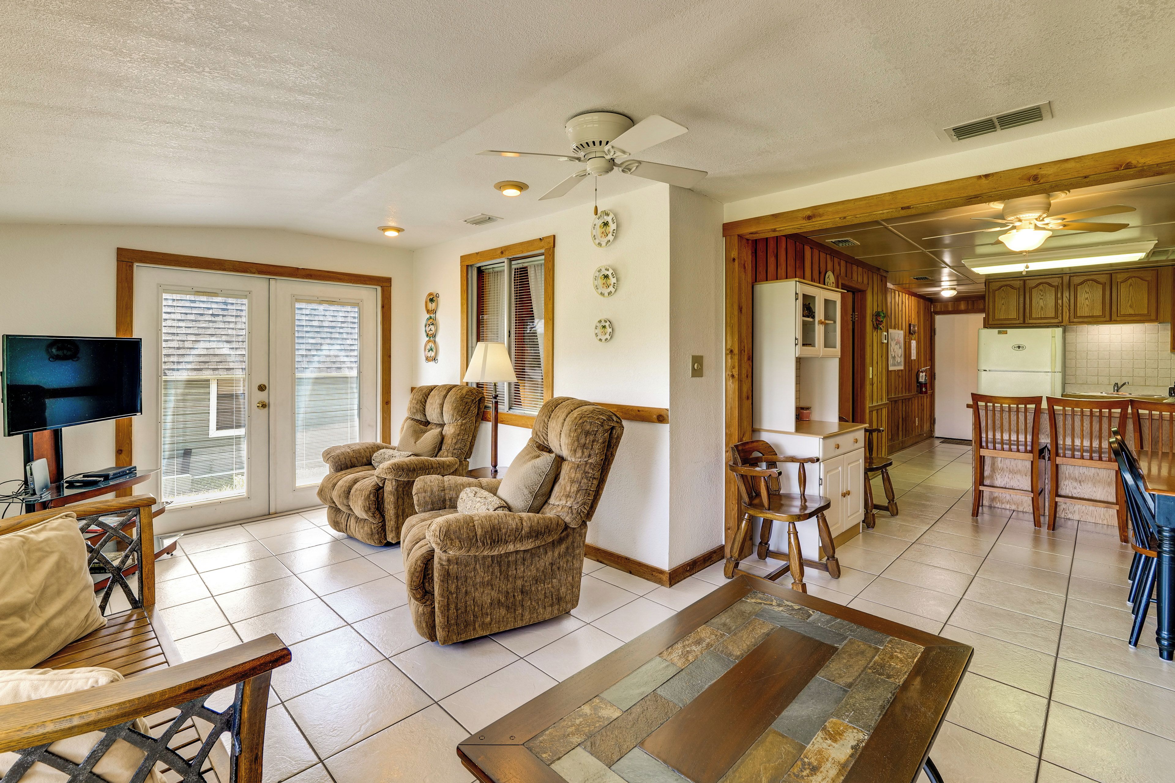 A spacious living area with a television, two armchairs, a coffee table, and a dining area with wooden chairs and a kitchen in the background.