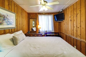 A room with wooden walls, a bed with white bedding, a ceiling fan, a television mounted on the wall, and a painting on the wall. - Coconut Cottage: Community Amenities, Near Disney (Clermont)