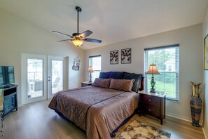 Une chambre à coucher comprenant un lit, un ventilateur de plafond, deux fenêtres avec stores, un téléviseur et un vase décoratif.