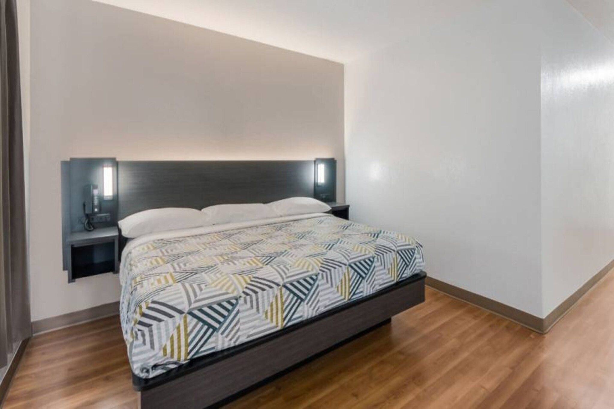 Une chambre d’hôtel moderne, équipée d’un lit, d’une table de chevet et d’un luminaire fixé au mur.