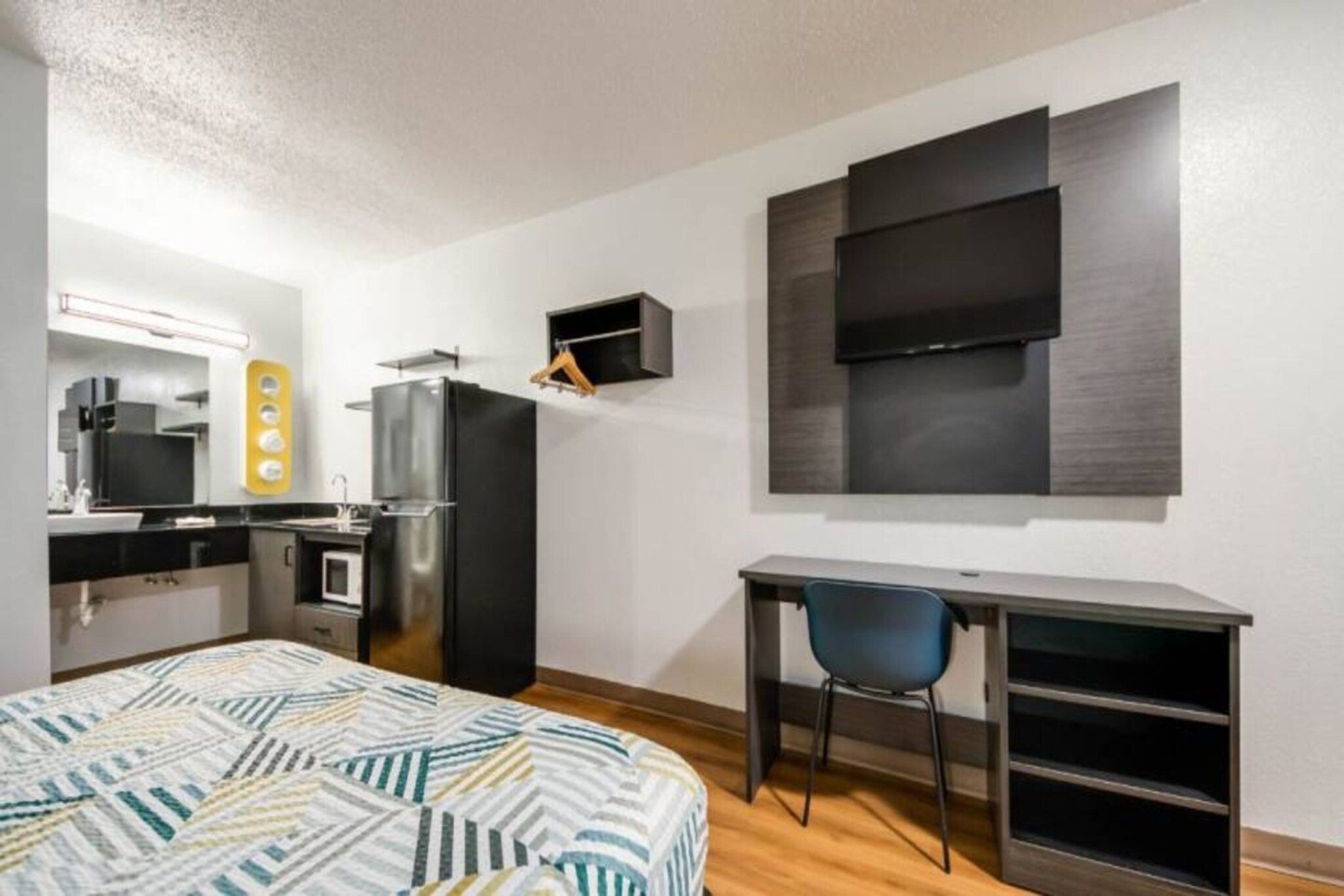 Quarto de hotel compacto com cama, escrivaninha, cadeira, cozinha compacta e televisão de tela plana fixada na parede.