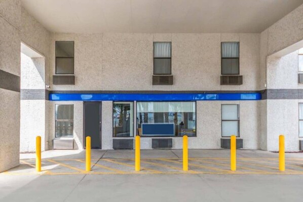 Entrada de un edificio con toldo azul, zona de estacionamiento con separadores amarillos y ventanas con persianas.