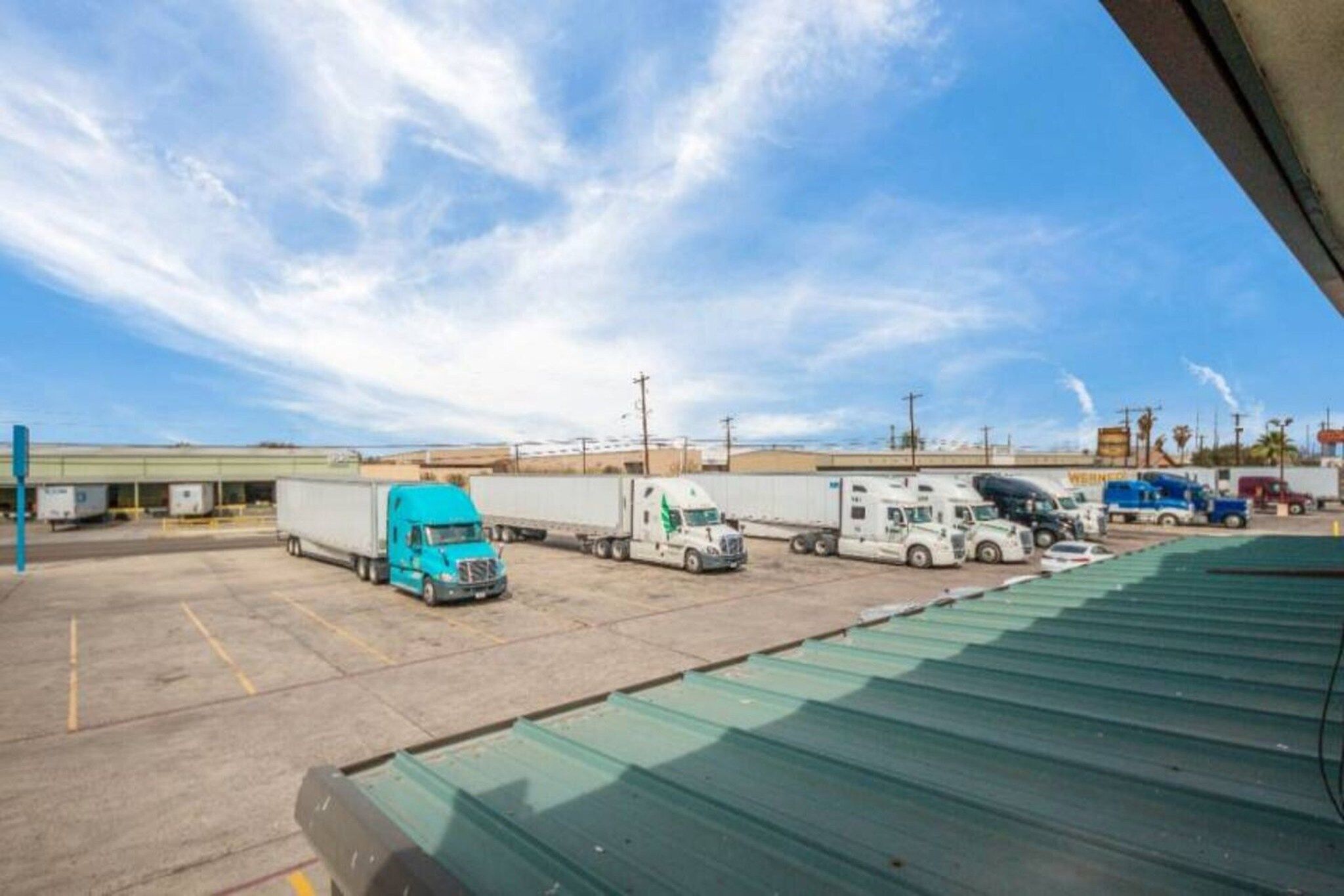 Een parkeerplaats met verschillende vrachtwagens en aanhangwagens, een heldere lucht en een gebouw op de achtergrond.