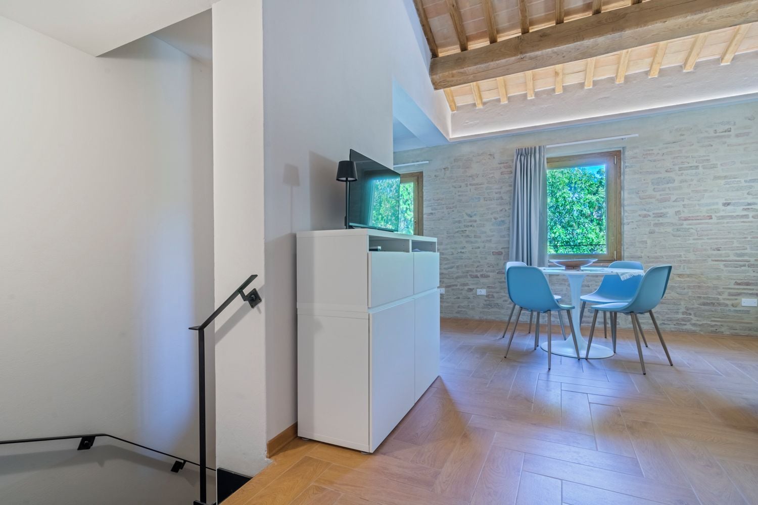 A modern kitchen with white cabinets, a black countertop, and a built-in TV. There is a dining area with a table and chairs, and a window with a view of greenery.