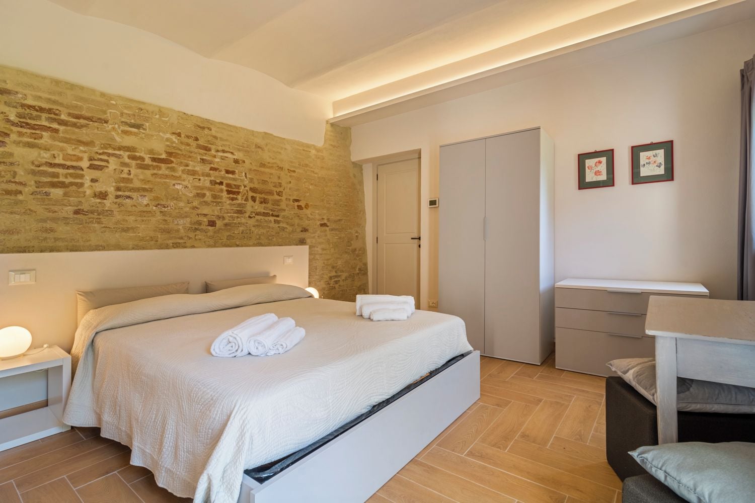 Un dormitorio con pared de ladrillo, cama con ropa blanca, mesita de noche con lámpara, armario y un pequeño escritorio con silla.